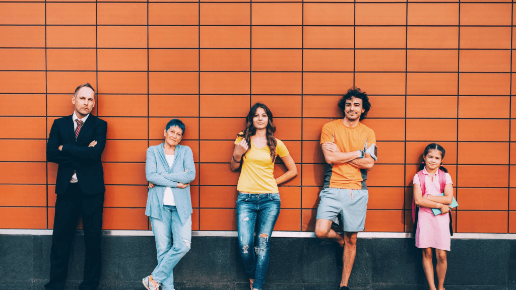 Five people representing different generations—from Baby Boomers to Gen Alpha—standing in a row against an orange tiled wall, illustrating demographic diversity.