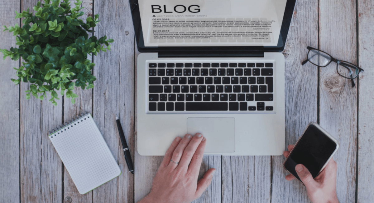 An overhead shot of a clean wooden desk featuring a laptop displaying a blog interface, a notebook, a pen, glasses, a smartphone, and a potted plant.