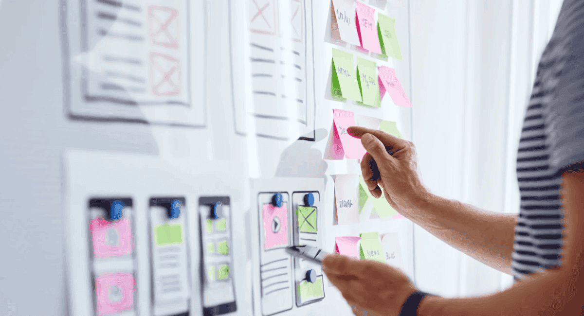 A person planning on a whiteboard covered with sketched app wireframes and colorful sticky notes, pointing at a pink sticky note while holding a smartphone.