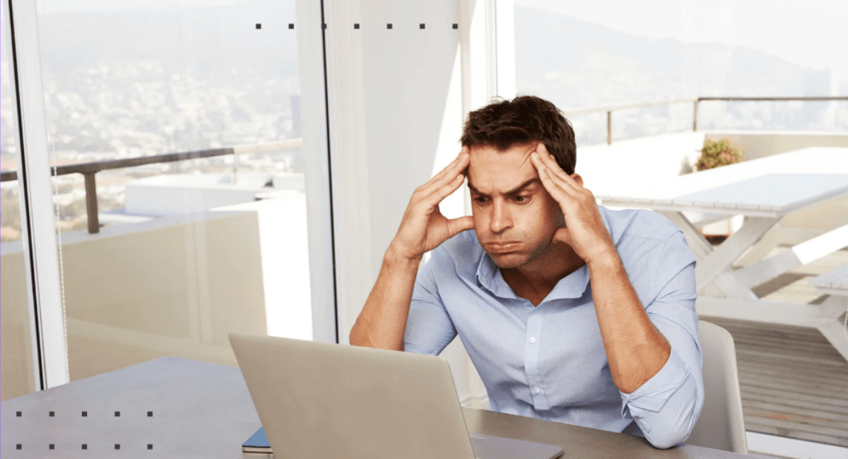 A stressed man sitting at a desk in a bright room, looking in frustration at his laptop screen with his hands pressing against his temples.
