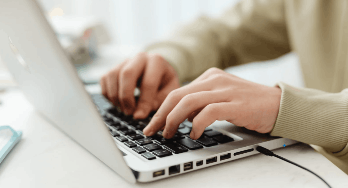 A close-up side view of hands typing on a laptop. The person is wearing a beige sweater, and a charging cable is plugged into the side of the device.