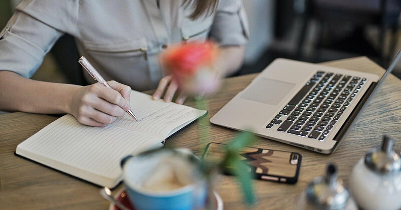 Woman working on laptop, writing notes in a notebook.