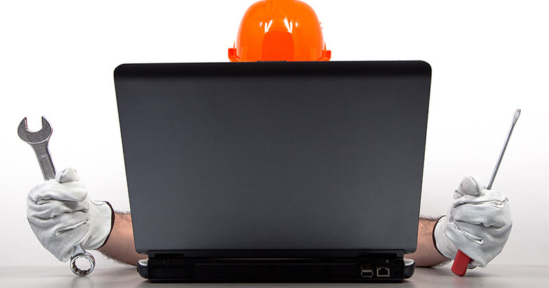 Professional wearing hard hat and gloves holding wrench and screwdriver working on laptop.