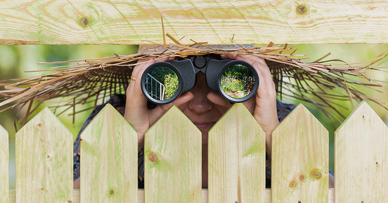 Person in straw hat hiding behind a fence looking through binoculars.