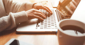 Close-up of a person in a light sweater typing on a laptop at a wooden table, with a cup of coffee and smartphone nearby.
