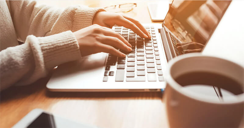 Close-up of a person in a light sweater typing on a laptop at a wooden table, with a cup of coffee and smartphone nearby.