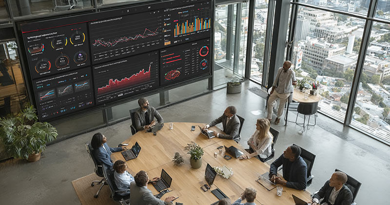 High-angle view of a modern corporate boardroom where a diverse group of executives is engaged in a strategic meeting. One executive stands and points to a large screen displaying SEO dashboards and performance analytics.