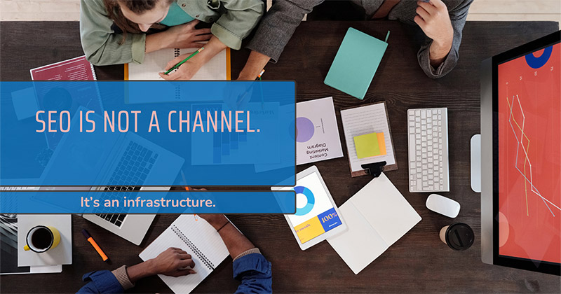 Overhead view of a wooden office table with digital devices, notebooks, printed charts, a coffee cup, and several people collaborating. A large translucent blue text overlay reads "SEO IS NOT A CHANNEL." followed by a smaller banner that states "It's an infrastructure."