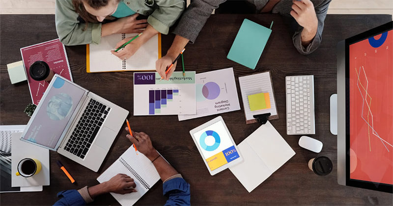 Overhead view of a wooden office table with digital devices, notebooks, printed charts, a coffee cup, and several people collaborating. A large translucent blue text overlay reads "SEO IS NOT A CHANNEL." followed by a smaller banner that states "It's an infrastructure."