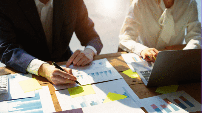 Two people sitting at a wooden desk reviewing printed charts and graphs. One person in a dark suit writes notes with a pen, while the other, wearing a white blouse, types on a laptop. Sunlight shines across the workspace scattered with papers and sticky notes.