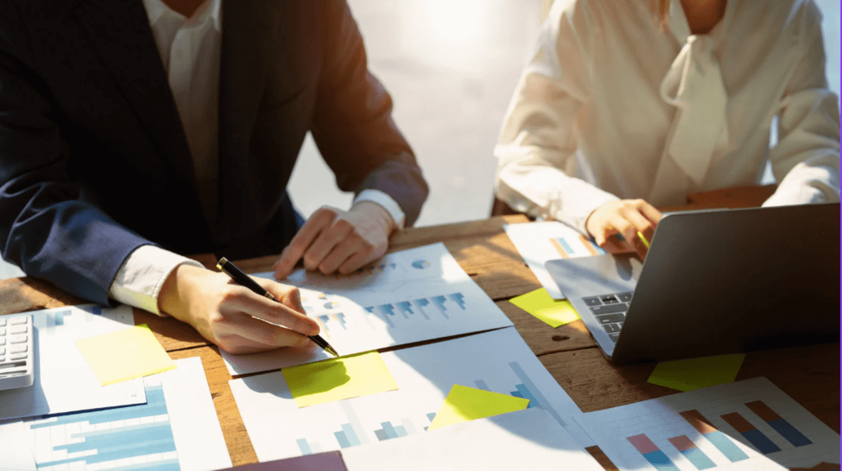 Two people sitting at a wooden desk reviewing printed charts and graphs. One person in a dark suit writes notes with a pen, while the other, wearing a white blouse, types on a laptop. Sunlight shines across the workspace scattered with papers and sticky notes.