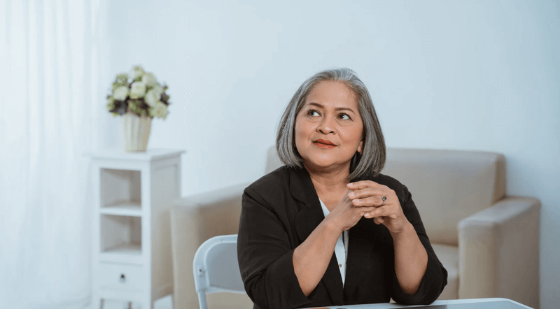 A confident middle-aged woman with gray hair sits at a table with her hands clasped, wearing a black blazer over a white top. She looks upward with a thoughtful expression in a bright, minimal office with beige furniture and a vase of flowers in the background.