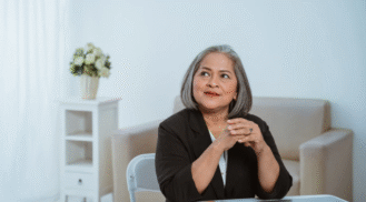 A confident middle-aged woman with gray hair sits at a table with her hands clasped, wearing a black blazer over a white top. She looks upward with a thoughtful expression in a bright, minimal office with beige furniture and a vase of flowers in the background.