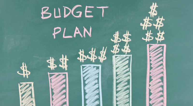 Green chalkboard illustration with the words “Budget Plan” written in pastel chalk above five bar graphs drawn in pink, blue, and green, each topped with increasing stacks of dollar signs to represent rising financial growth.
