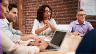 A diverse group of four professionals sits around a conference table in a modern office with exposed brick walls. A woman at the center speaks while others listen attentively, laptops and notebooks open in front of them.