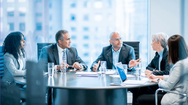 Executives sitting around a round conference table in a modern office, discussing documents and laptops during a business meeting.
