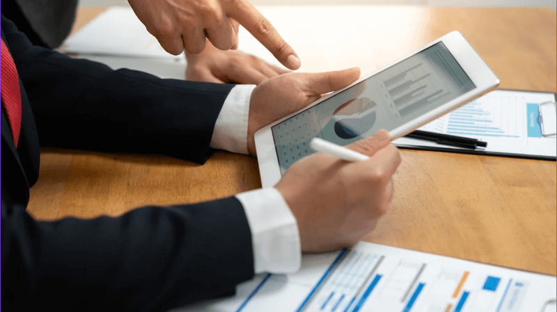 Close-up of two people reviewing charts and graphs on a tablet, with hands pointing at data and printed reports on the table