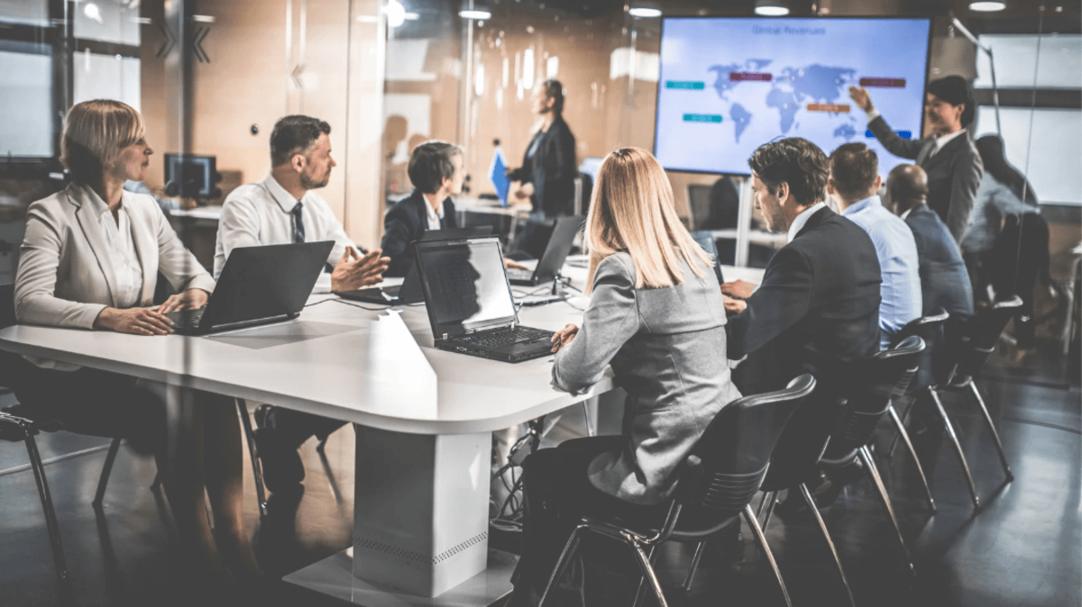 Business professionals seated at a long white conference table with laptops while a presenter points to a world map on a screen at the front of the room.
