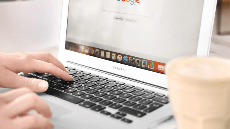 Hands typing on a MacBook Air with the Google homepage displayed on the screen, with a blurred coffee cup in the foreground.