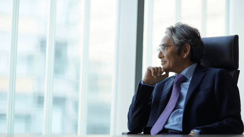 A senior professional man in a suit and tie sitting at a desk, looking thoughtfully out of large office windows.