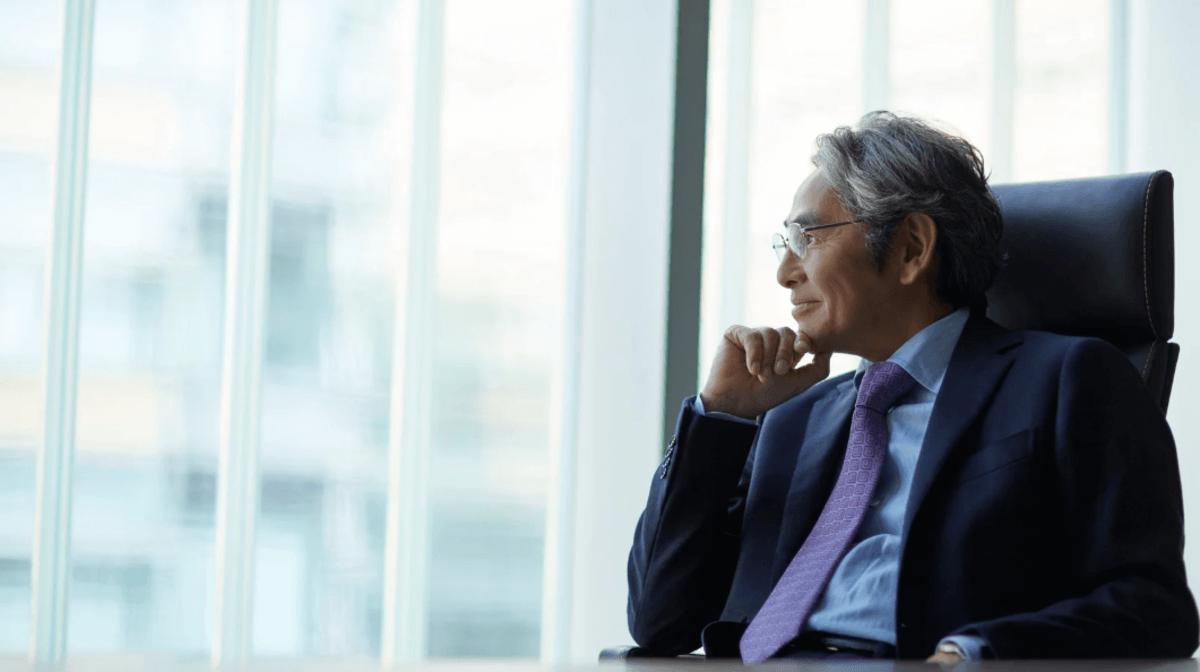 A senior professional man in a suit and tie sitting at a desk, looking thoughtfully out of large office windows.