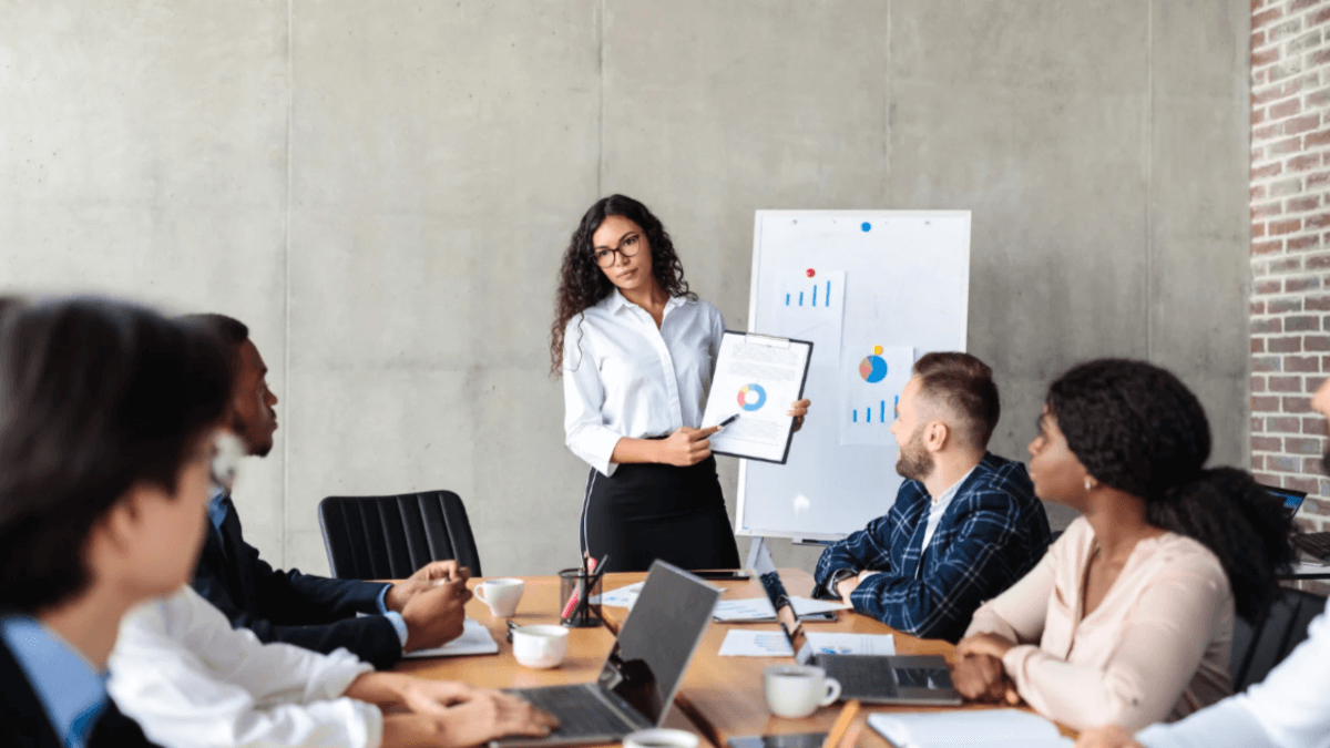 Businesswoman presenting charts to a group of colleagues in a conference room, pointing to data on a clipboard and whiteboard during a team meeting.