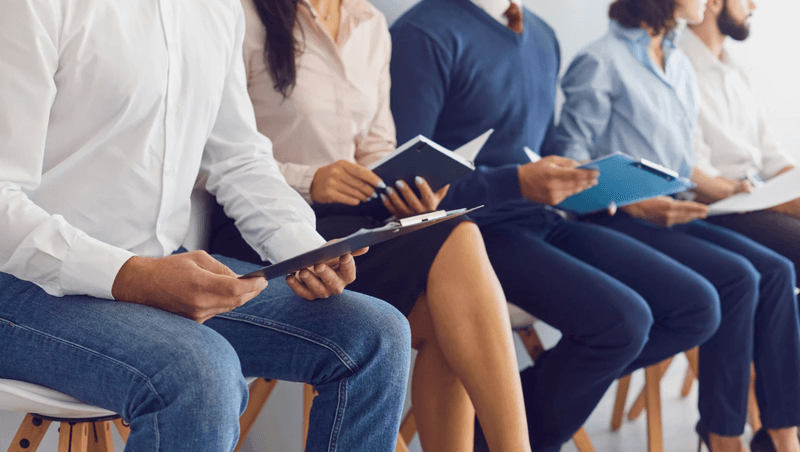 Group of job applicants sitting in a row holding resumes during a hiring process, representing competitive SEO job market conditions.