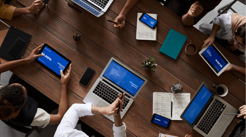 Overhead view of a marketing team collaborating at a conference table using laptops, tablets, and smartphones to review digital strategy and analytics.