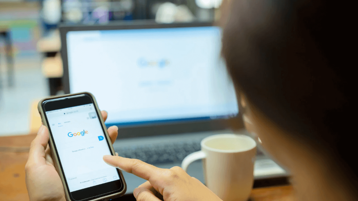 Person using a smartphone to search Google with a laptop in the background, illustrating mobile and desktop search behavior.