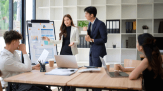 Business team in a conference room as two presenters stand beside a flip chart with charts and graphs, explaining data to three colleagues seated at a table with laptops and coffee cups.