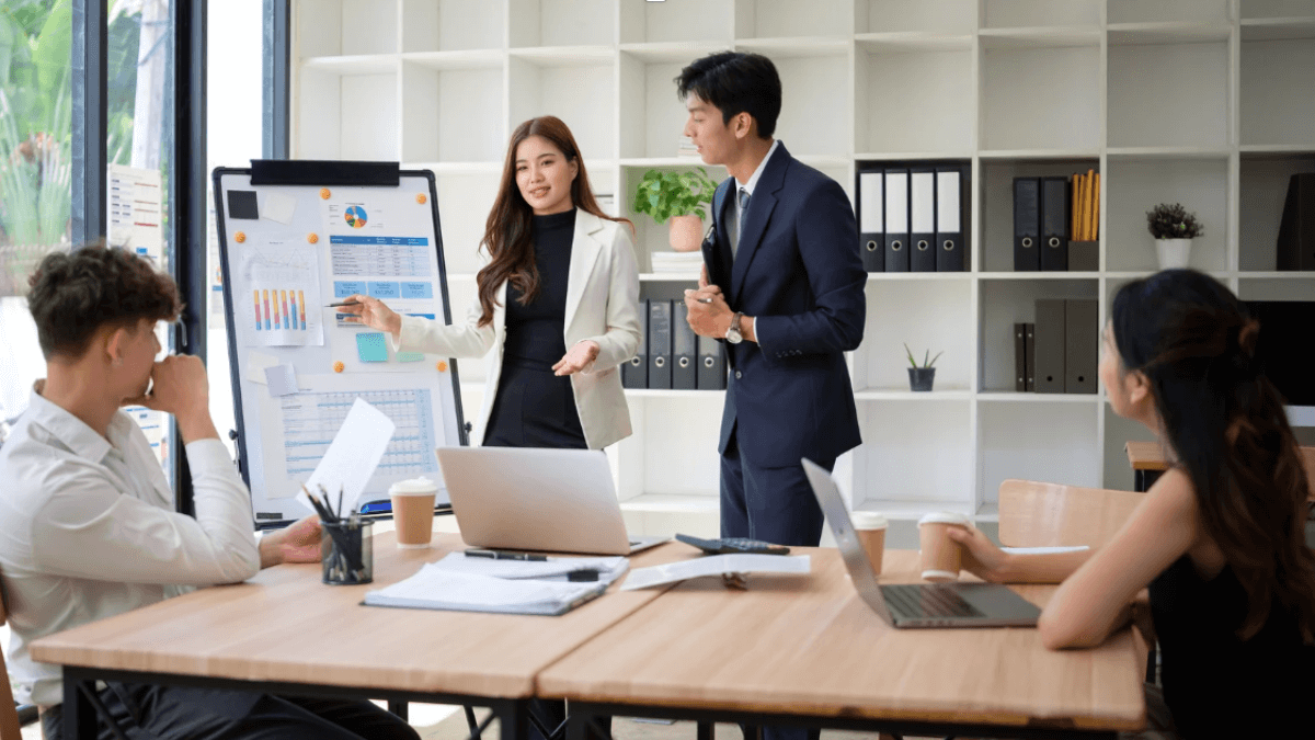 Business team in a conference room as two presenters stand beside a flip chart with charts and graphs, explaining data to three colleagues seated at a table with laptops and coffee cups.