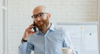 Man talking on a phone while holding a coffee in a bright office with charts on a whiteboard behind him
