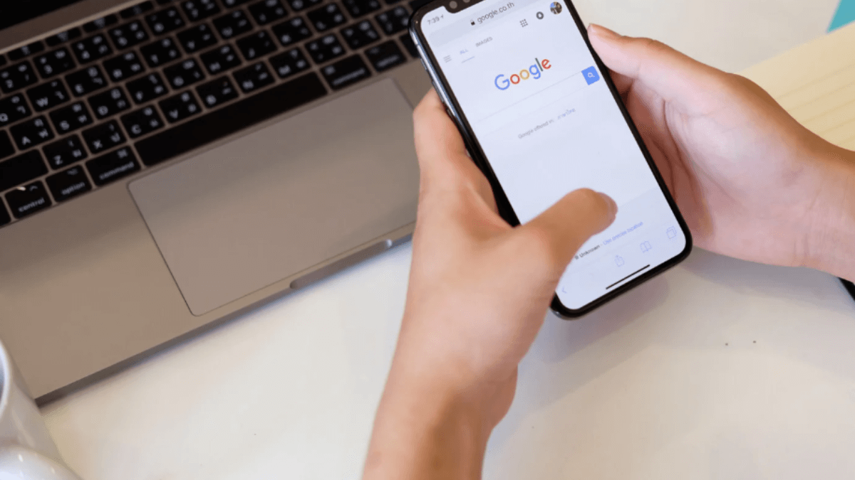 Person using a smartphone with Google search open beside a laptop, representing mobile search behavior and cross-device browsing.