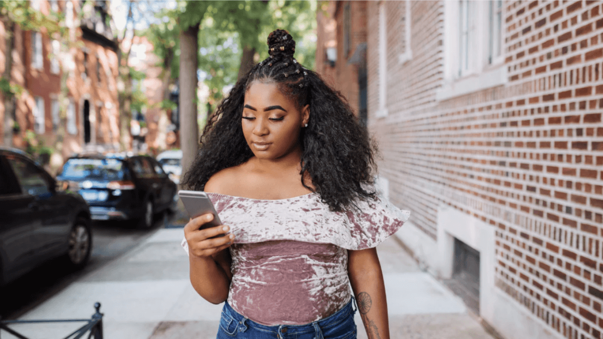 Young woman walking down a city sidewalk while checking her smartphone, illustrating mobile search and on-the-go information access.