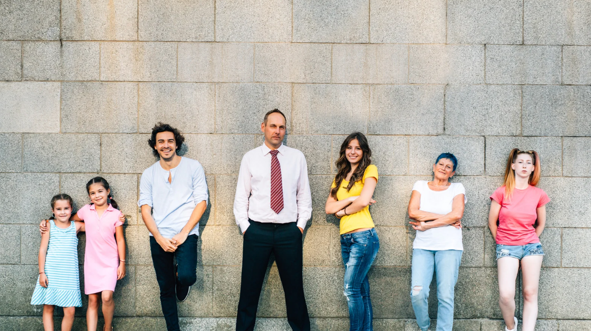 A group of six people of various ages standing casually against a grey stone wall, representing a multi-generational family or demographic cross-section for market research.