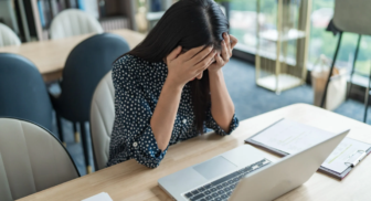 Stressed woman holding her head while working on a laptop with paperwork at a table