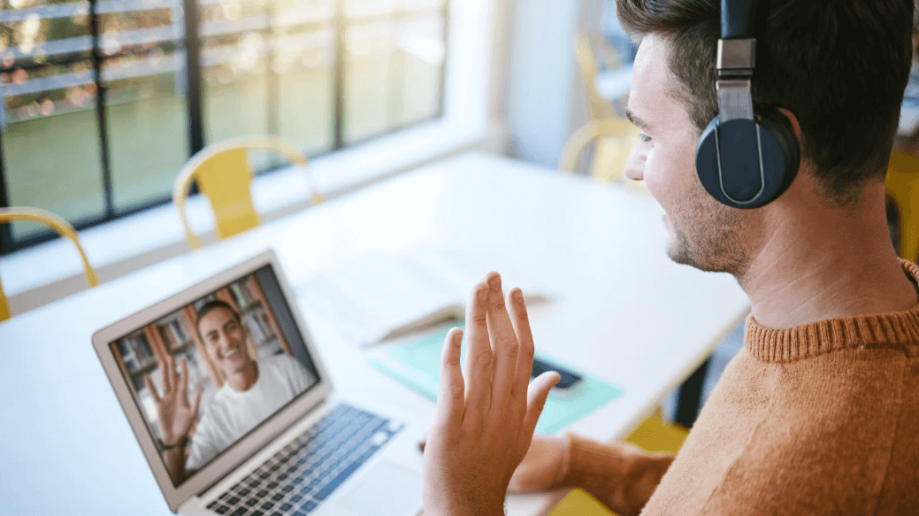 Remote business meeting with two professionals on a video call, one wearing headphones and waving at a colleague on a laptop screen