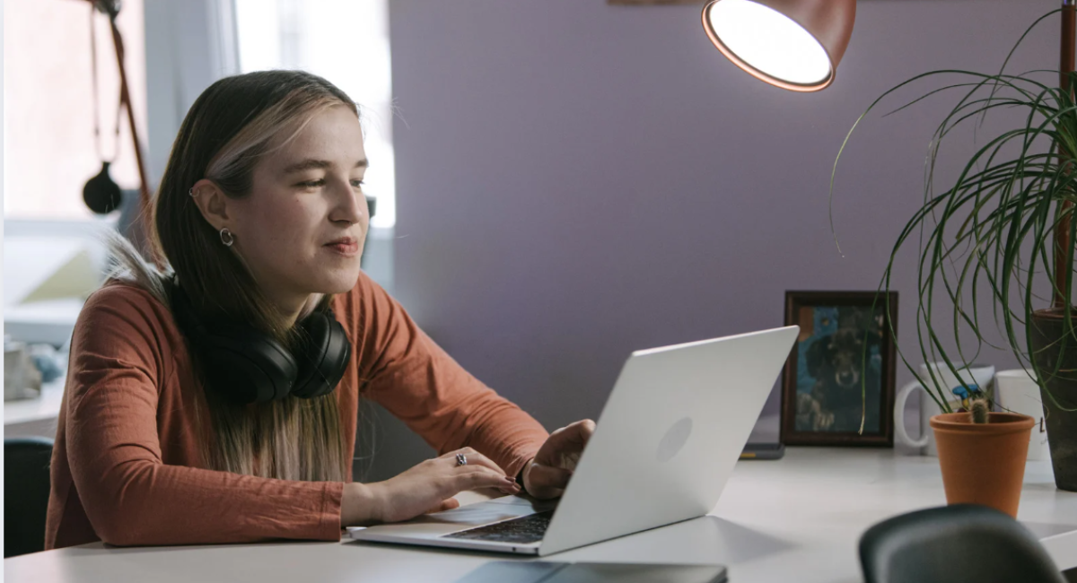 Woman working on a laptop with headphones at a home office desk under a reading lamp