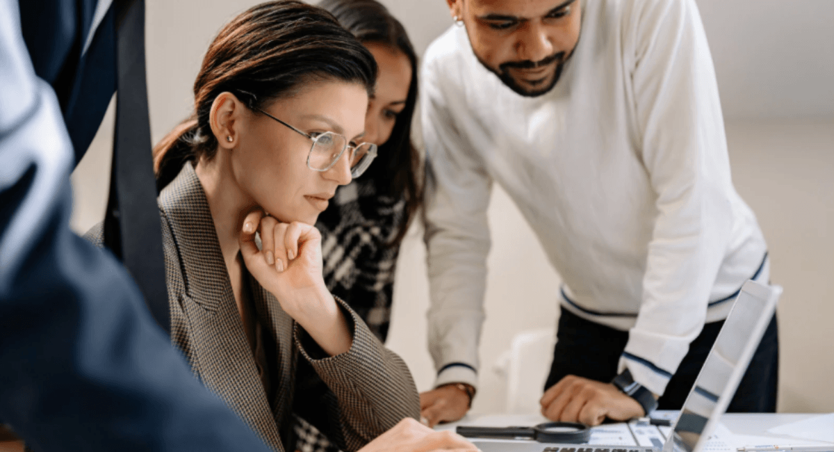 Three business professionals leaning in and looking intently at a laptop screen together in an office setting.