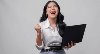 A joyful woman in a white button-down shirt cheering with her fist raised in celebration while holding an open laptop.