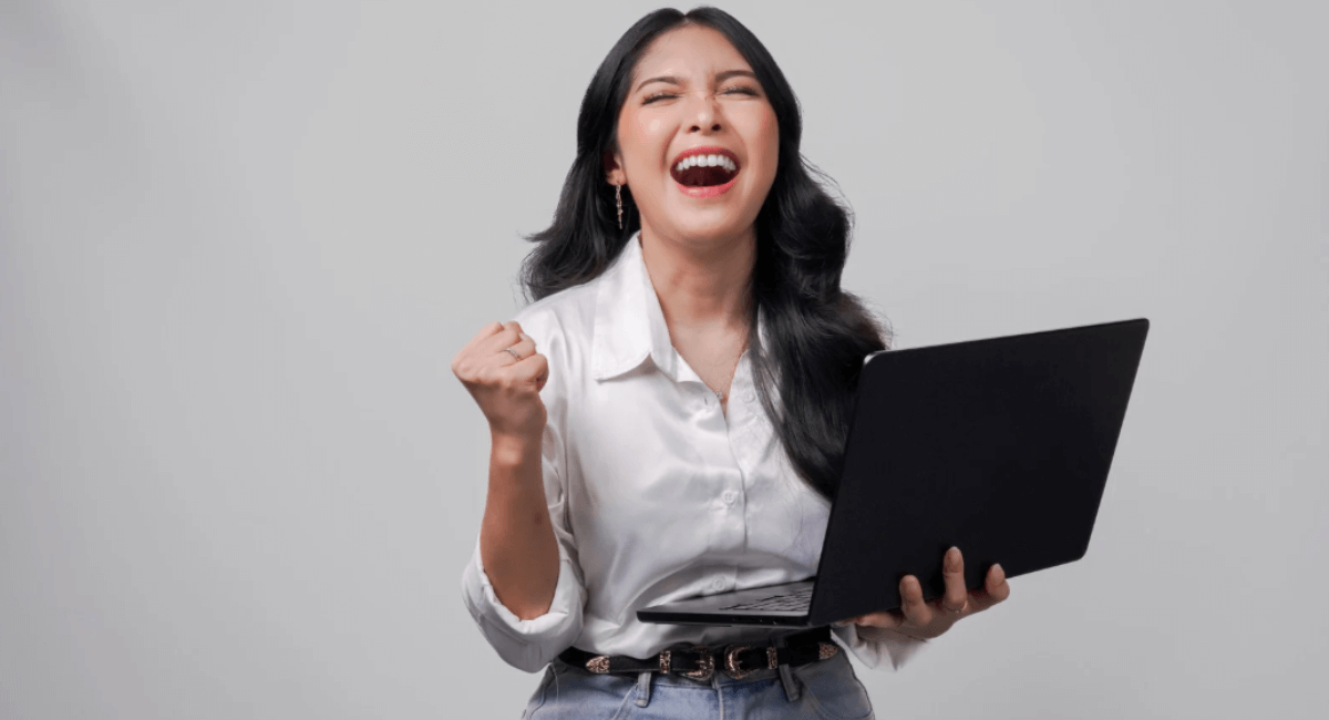 A joyful woman in a white button-down shirt cheering with her fist raised in celebration while holding an open laptop.