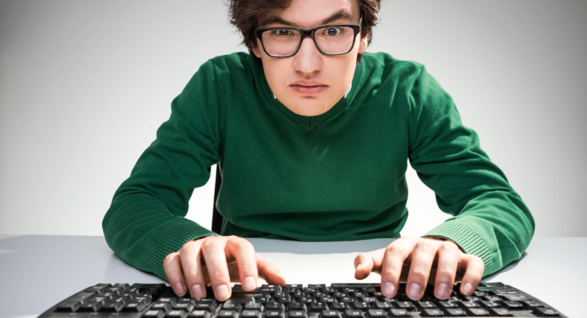 A young man with glasses and a green sweater leaning forward and typing intensely on a black computer keyboard.