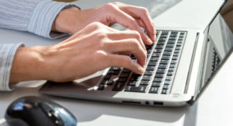 Close-up of a person's hands typing on a silver laptop keyboard next to a black wireless computer mouse.
