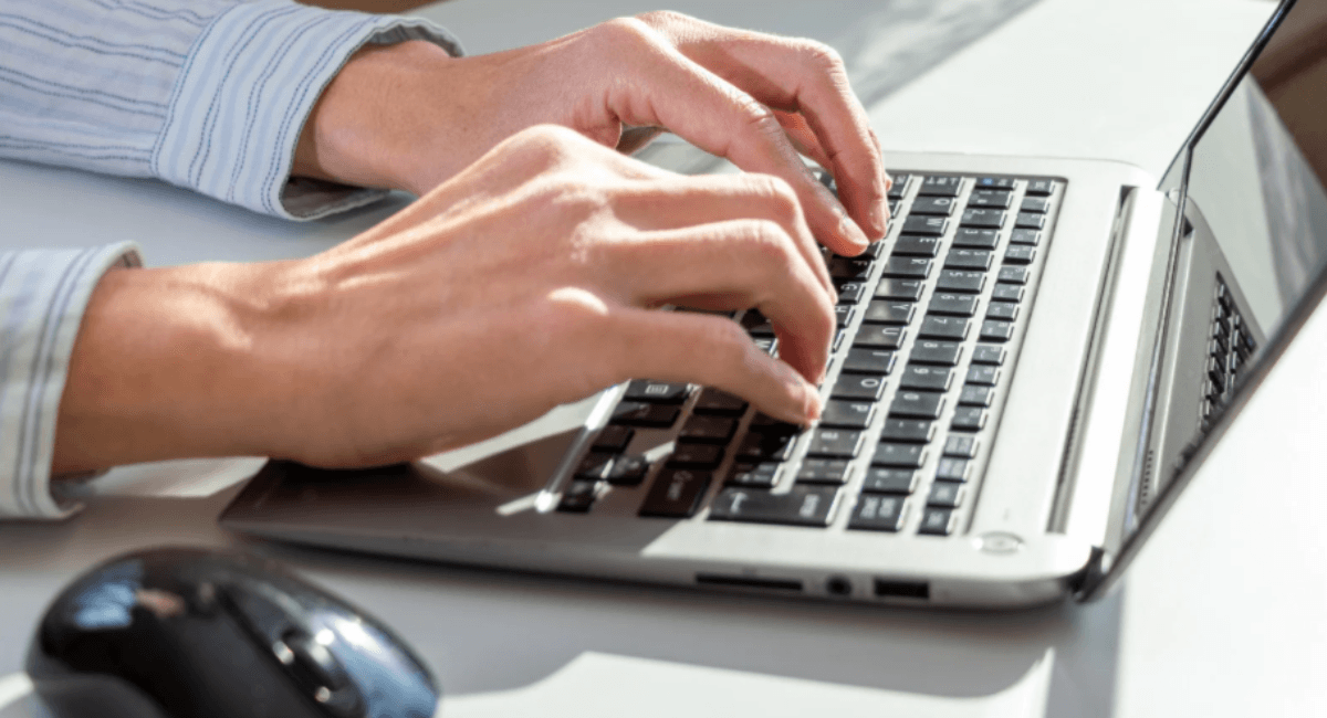 Close-up of a person's hands typing on a silver laptop keyboard next to a black wireless computer mouse.