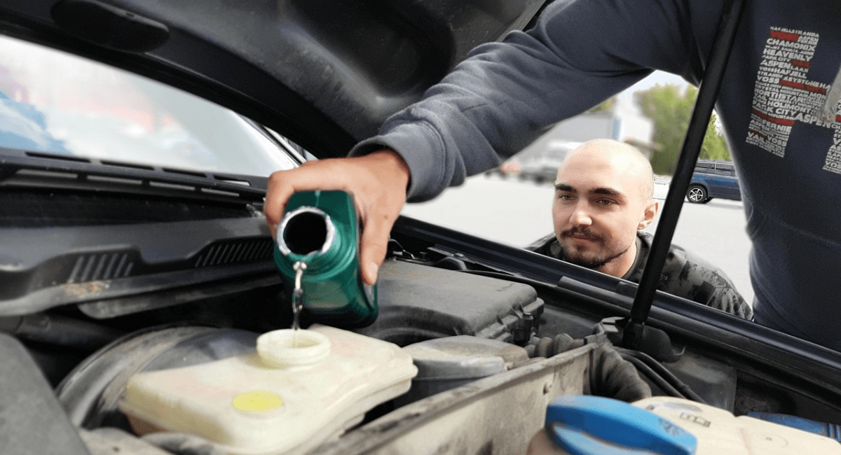 A close-up shot of a person pouring fluid from a green bottle into a car's fluid reservoir under the open hood, while another man watches closely in the background.