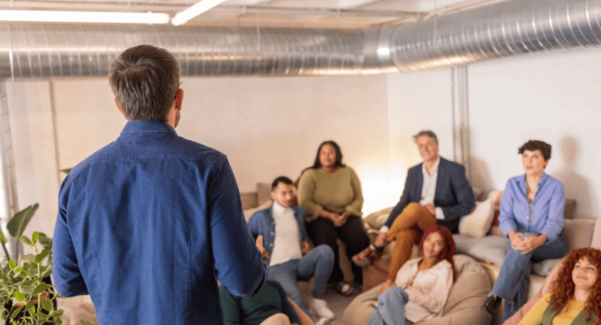 A presenter viewed from behind speaking to a diverse, attentive audience seated comfortably on beanbag chairs in a modern office space.