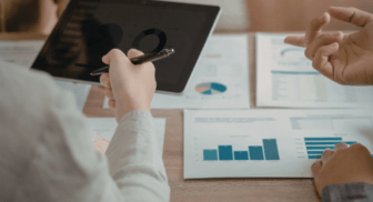 A business team collaborating at a table, pointing at printed data charts and a tablet displaying various performance graphs and pie charts.