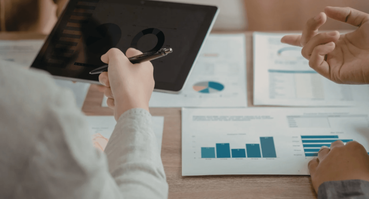 A business team collaborating at a table, pointing at printed data charts and a tablet displaying various performance graphs and pie charts.