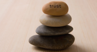 A stack of four smooth balancing stones resting on a wooden surface, with the top stone featuring the word "trust" written on it.