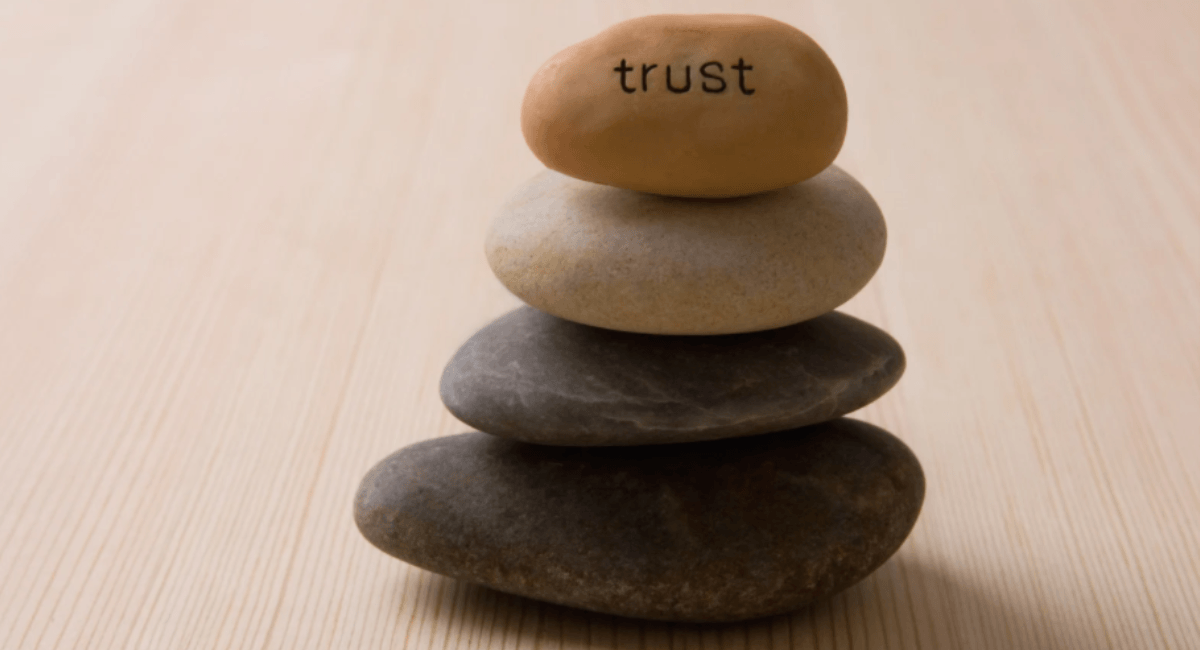 A stack of four smooth balancing stones resting on a wooden surface, with the top stone featuring the word "trust" written on it.